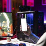 A gamer wearing a headset sits at a desk with a PC, holding up a smartphone with a blank white screen. A colorful first-person shooter game is displayed on the monitor, with neon lighting and a glass-sided gaming PC glowing in the background.