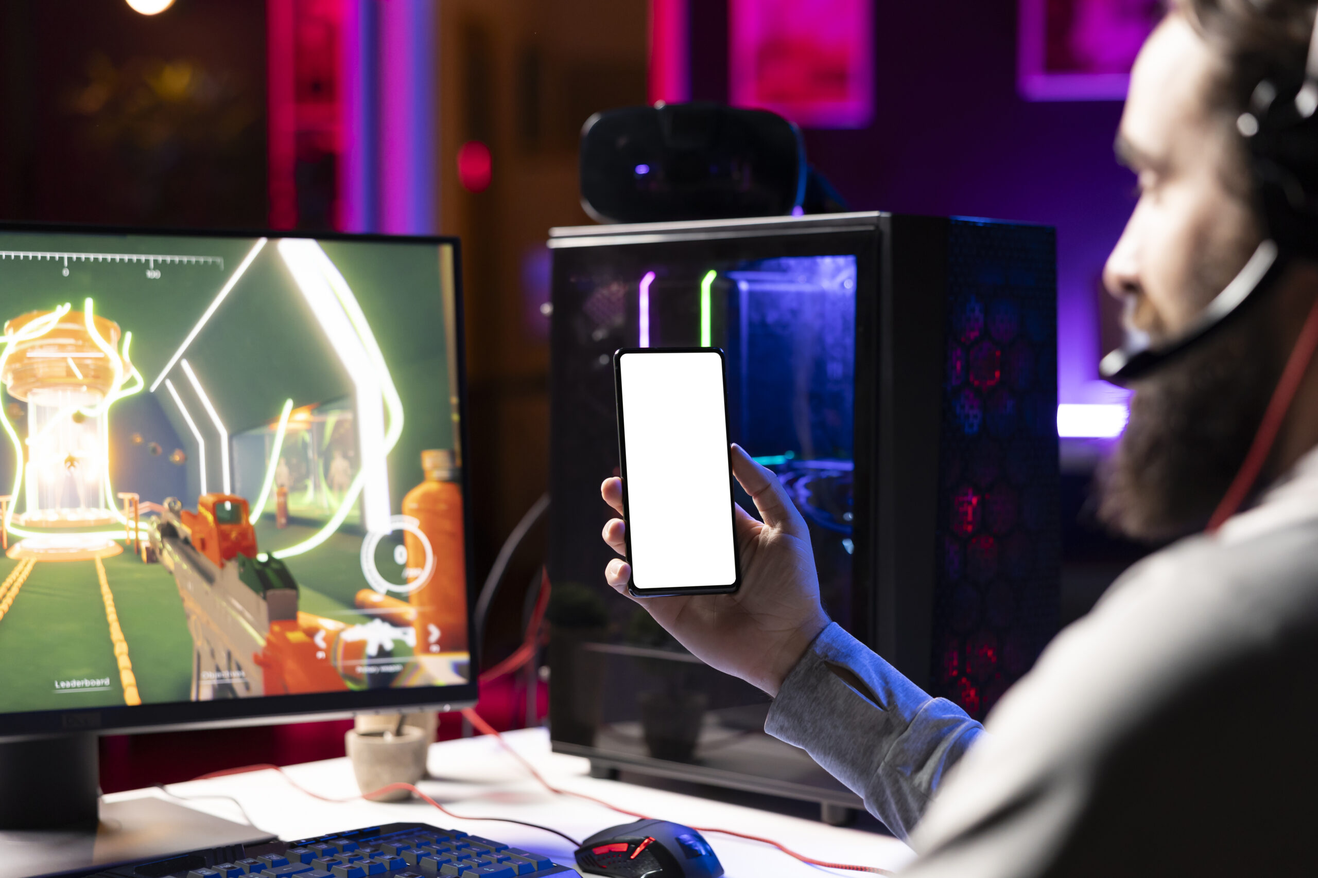 A gamer wearing a headset sits at a desk with a PC, holding up a smartphone with a blank white screen. A colorful first-person shooter game is displayed on the monitor, with neon lighting and a glass-sided gaming PC glowing in the background.